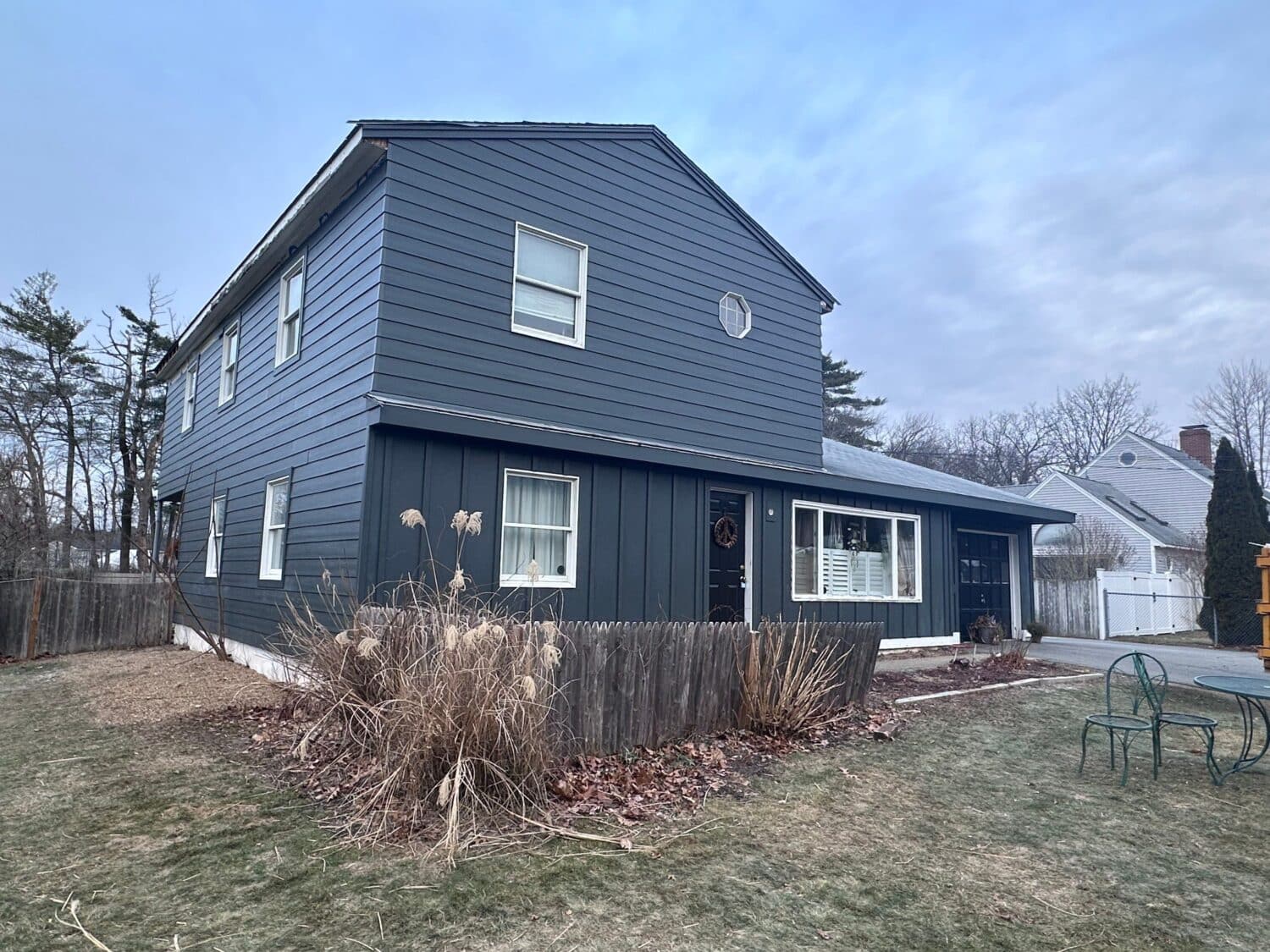 A two-story dark gray house with white window frames, a small circular window above, and a dry plant bed in front. The yard is partially fenced and bordered by winter-bare trees, with a cloudy sky above.