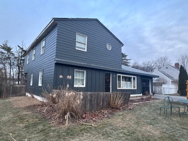 A two-story dark gray house with white window frames, a small circular window above, and a dry plant bed in front. The yard is partially fenced and bordered by winter-bare trees, with a cloudy sky above.