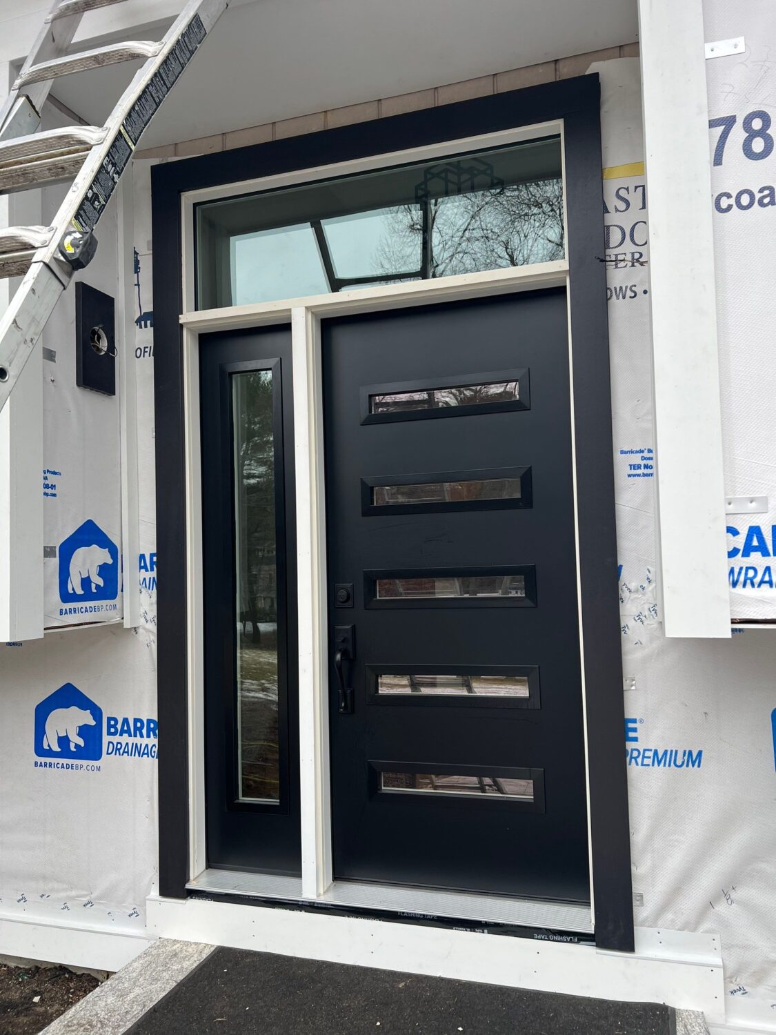 A modern black door with four horizontal glass panels is set in a white frame. A narrow vertical window is on the left side of the door. The exterior wall is partially covered with house wrap, and a ladder leans against the wall.
