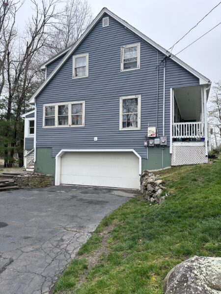 A gray two-story house with a white garage door and a small porch. The exterior features white-trimmed windows and green basement walls. The front yard has grass and a stone path. Trees are in the background.