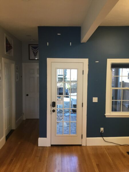 A white-framed glass door set in a blue wall leads to an external patio. The room has wooden floors and a window with blinds on the right. A hallway with closed white doors is visible on the left.