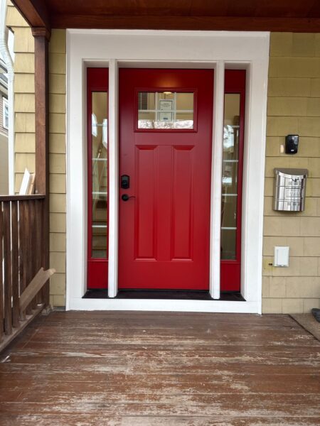 A red front door with two vertical glass panels on each side and a horizontal window at the top. The door has a black handle and is set against a light green house exterior. A metal mailbox is mounted on the right side of the entryway.