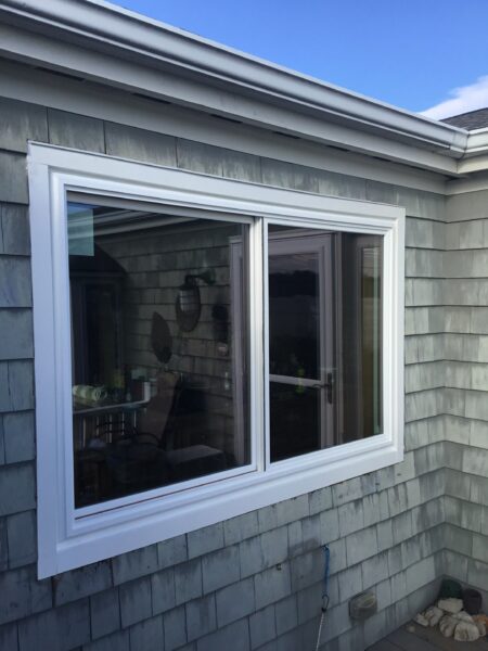 A white-framed window is set in a gray shingle-clad wall of a house. The window is sliding and reflects light. The sky is visible at the top, and some garden items are seen below the window.