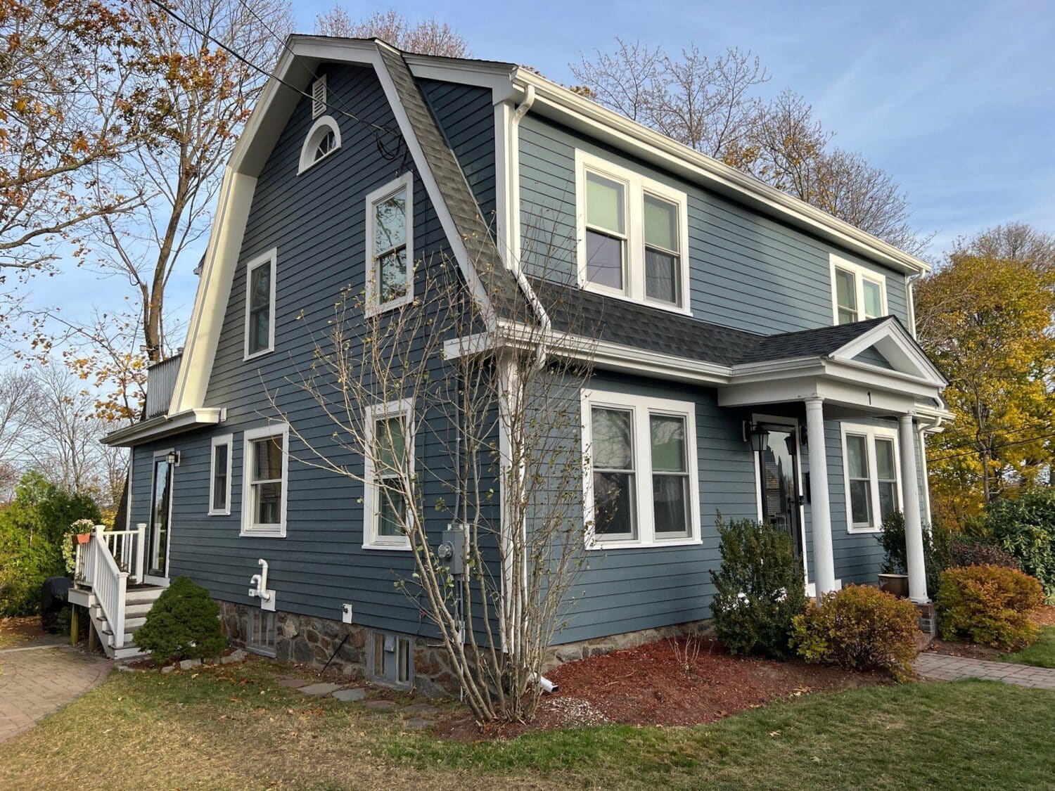 A two-story house with dark blue siding and white trim, featuring a gabled roof and four visible windows. A small porch with steps leads to the front door. The yard has sparse shrubs, and trees with autumn leaves surround the house.