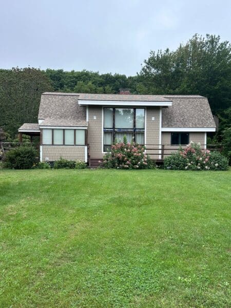 A small, single-story house with tan siding and a gabled roof. It has large windows, surrounded by shrubs and flowers. The house is set against a backdrop of dense, green trees, and theres a large, grassy field in the foreground.