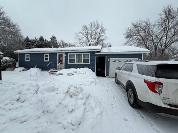 A blue single-story house with snow-covered roof and yard is shown. Snow is piled beside the driveway, where a white SUV is parked. Leafless trees are in the background under a cloudy, gray sky.