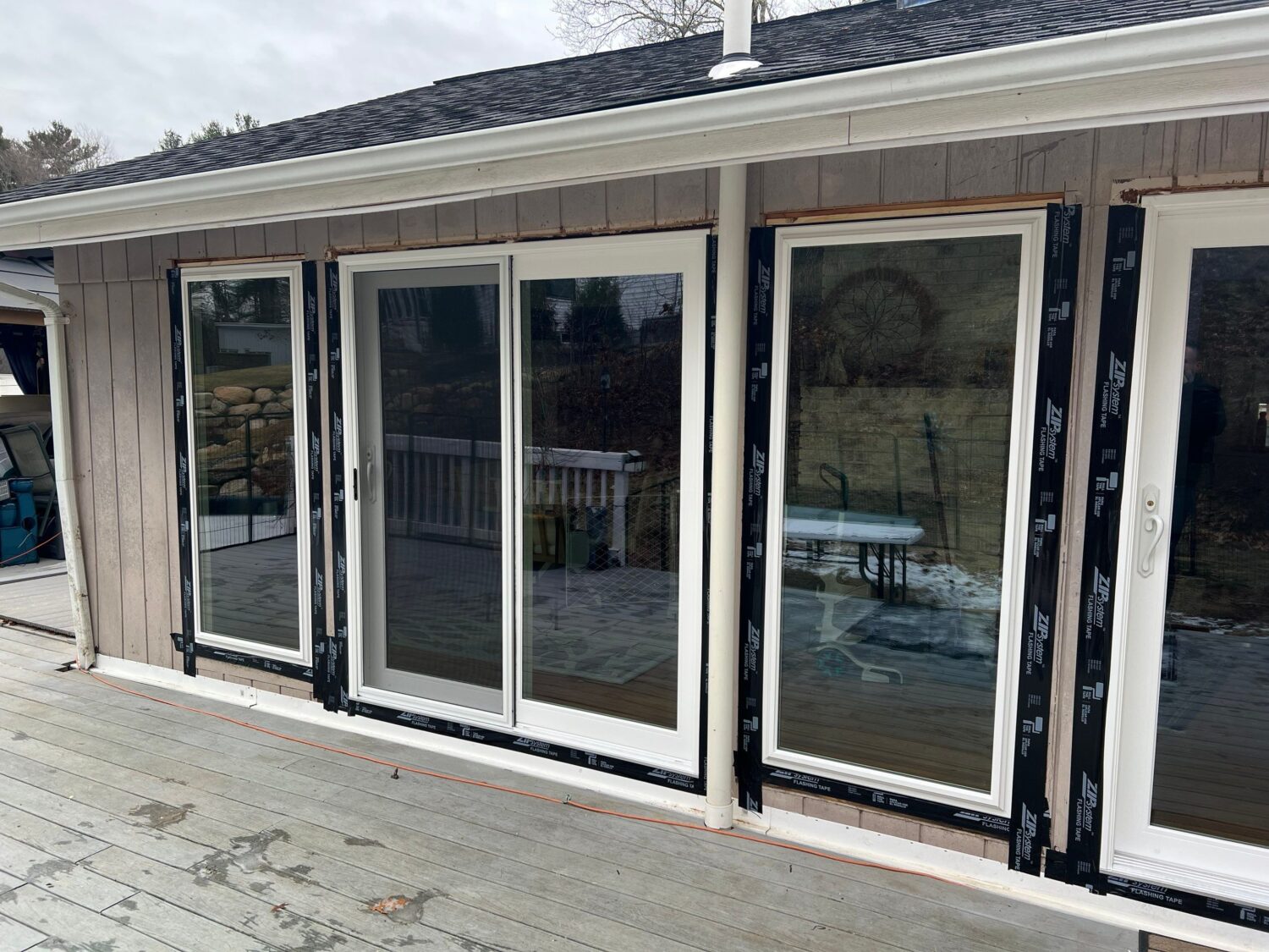 A house exterior with three sliding glass doors, newly installed with visible black insulation strips. The doors lead to a back deck, with a view of a backyard and patio furniture. Overcast sky in the background.