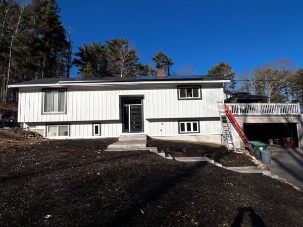 A white split-level house with a black front door and several windows. Theres a red ladder leaning against the right side, leading up to a deck. The yard has stone steps and fresh soil. The sky is clear blue, and trees are visible in the background.