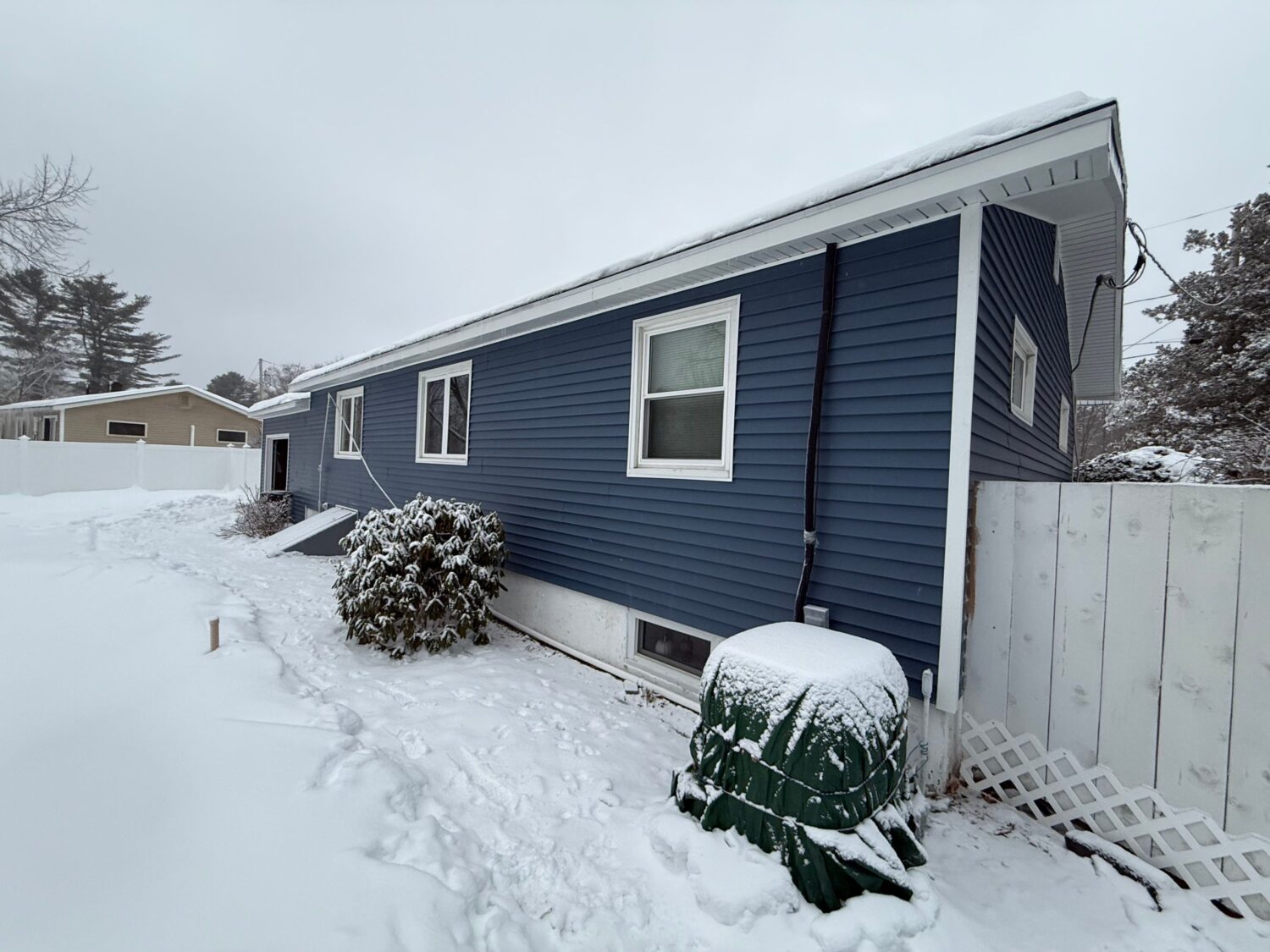 A blue house with snow-covered ground and roof. A bush near the house is partially covered in snow. There is a green object, possibly a utility cover, with snow on it. A white fence is on the right, and trees are visible in the background.