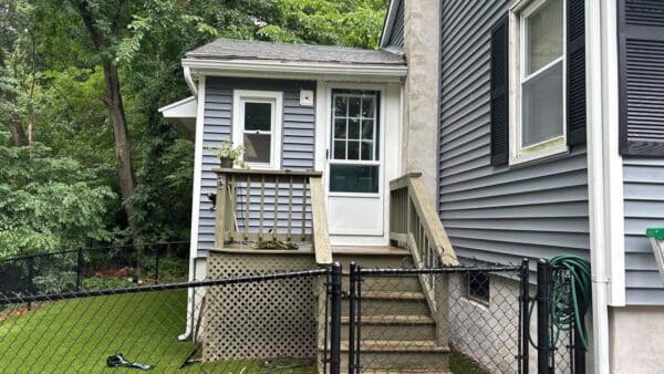 Small back porch of a house with gray siding, featuring a white door and steps leading down to a fenced yard with greenery. A hose is coiled on the side, and a small potted plant is on the porch railing.