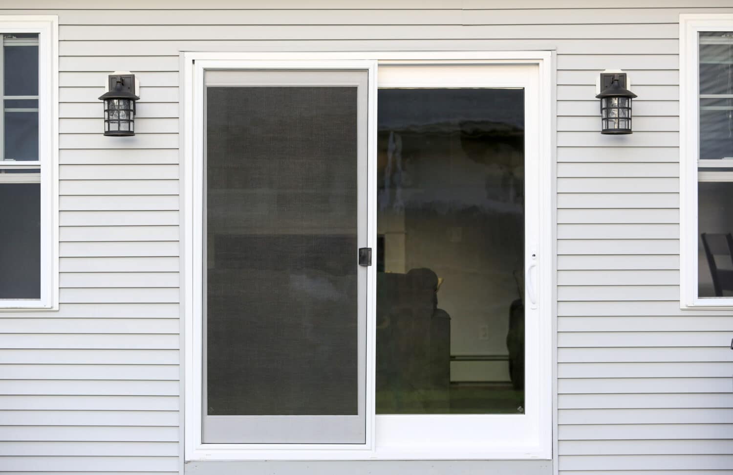 Sliding glass door on a gray vinyl-sided house with a screen on the left and clear glass on the right. Two black outdoor wall lanterns are mounted on each side of the door. Reflections can be seen on the glass.