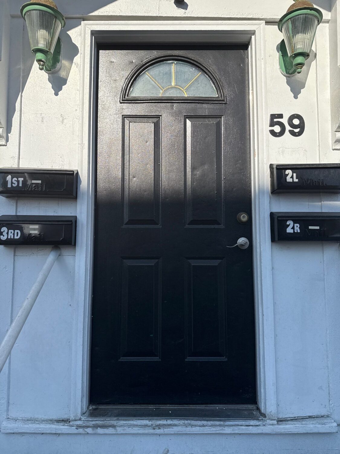 A black door with an arched window sits between four mailboxes labeled 1st, 2L, 2R, and 3rd. The door is flanked by two green and white lanterns, and the number 59 is displayed on the wall.