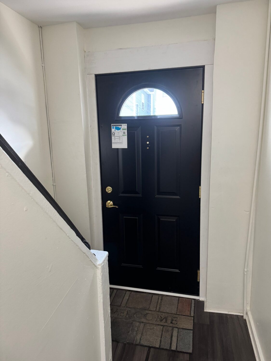 A black front door with a semicircular window at the top is seen from inside a hallway. Theres a staircase to the left and a multicolored welcome mat on the floor. A notice is attached to the door.