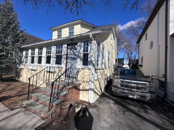 A two-story yellow house with white trim featuring a small porch and steps leading to the front door. A large tree casts shadows on the house. A silver pickup truck is parked in the driveway beside the house. Clear, sunny day with blue skies.