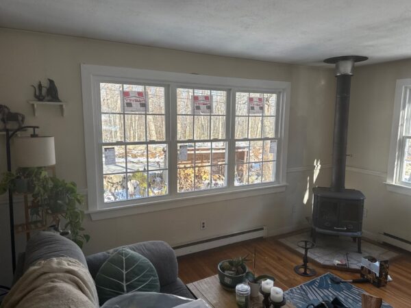 A cozy living room with a large window displaying a winter scene outside. Theres a wood-burning stove in one corner and a couch with a leaf-patterned pillow in the foreground. Various small items are on a wooden floor and a side table.