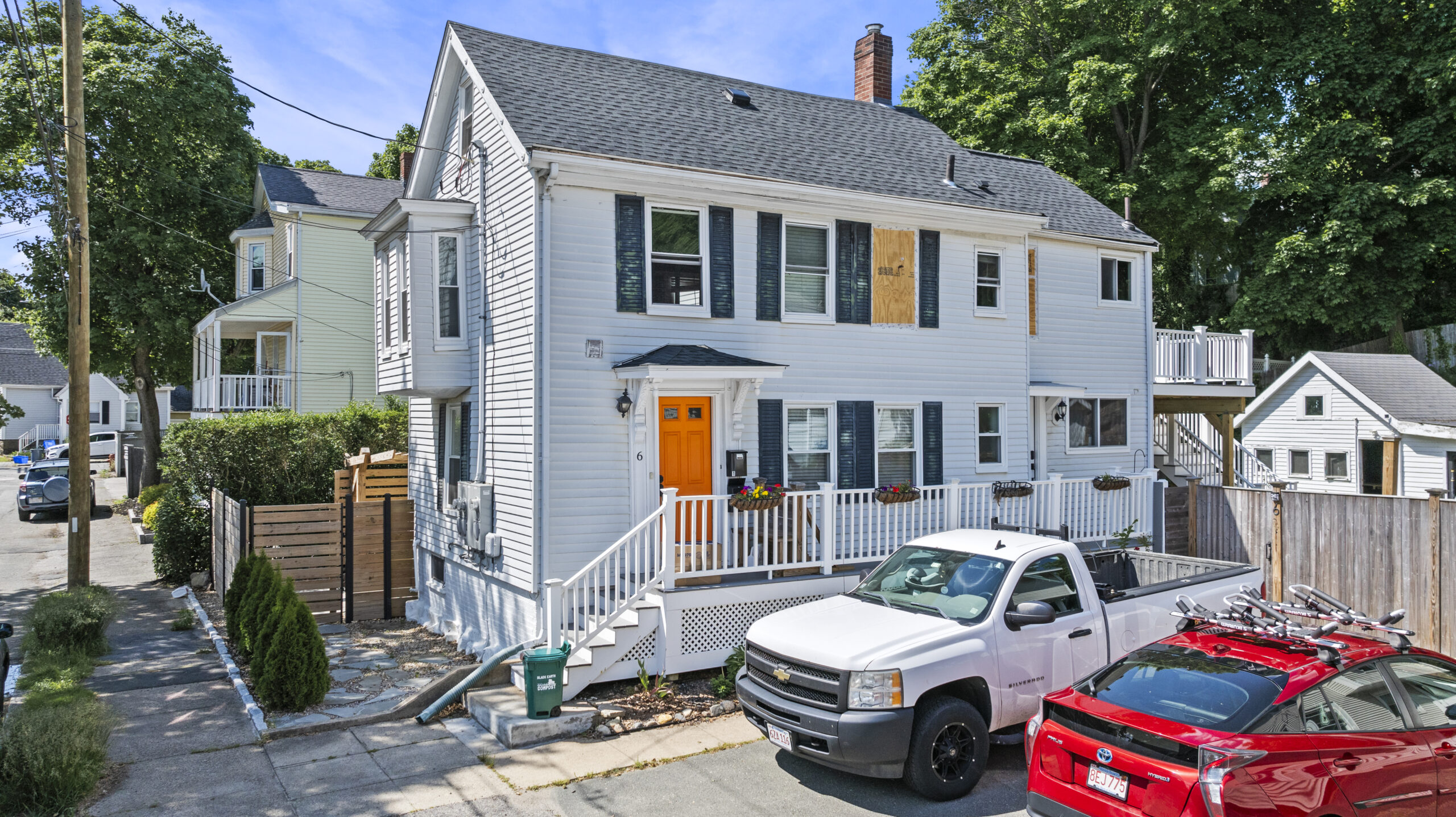 A two-story white house with James Hardie siding, a front porch, orange door, and flower boxes under the windows sits on a corner lot. A white pickup truck and red car are in the driveway, with trees in the background.