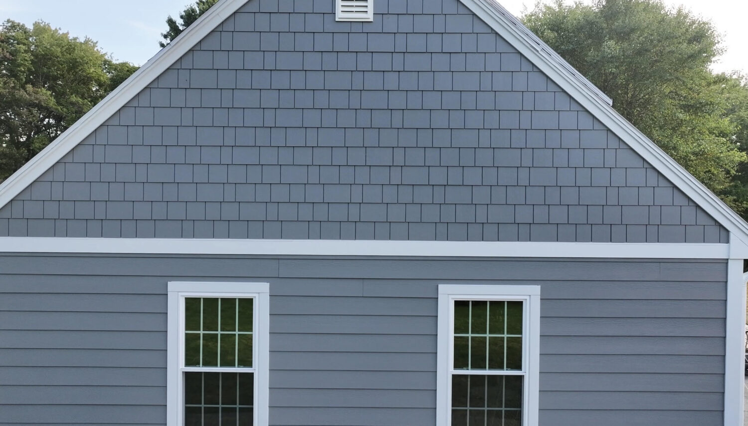A close-up view of the side of a gray house with two white-framed windows, gray siding and shingles, and green trees visible in the background.