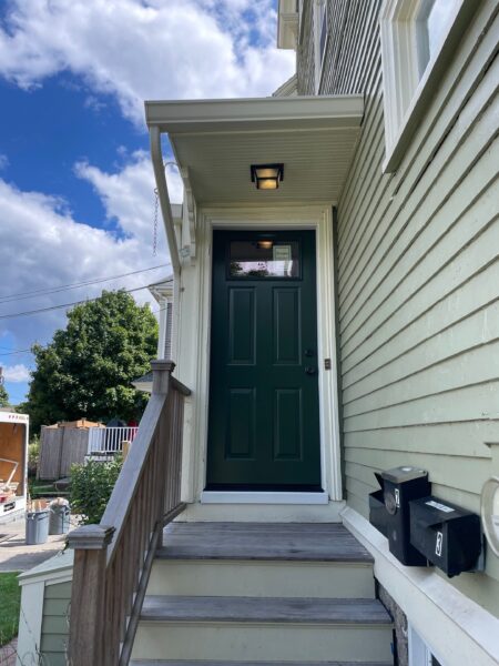 A green door with four panels at the top of a small wooden staircase, attached to a light green house. The door has an awning and outdoor light above, with two black mailboxes mounted nearby. The sky is partly cloudy.