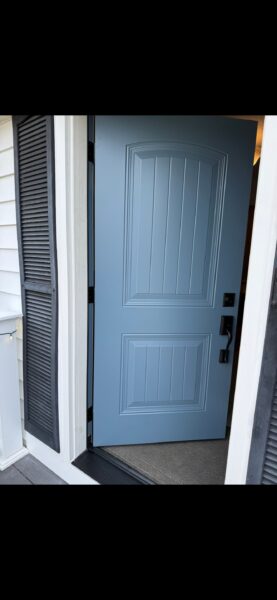 A light blue front door with decorative panels is partially open, revealing a carpeted entryway. The door is set in a white exterior wall with black shutters on either side.