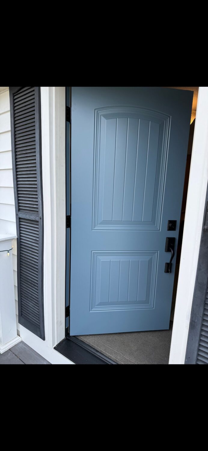 A light blue front door with decorative panels is partially open, revealing a carpeted entryway. The door is set in a white exterior wall with black shutters on either side.