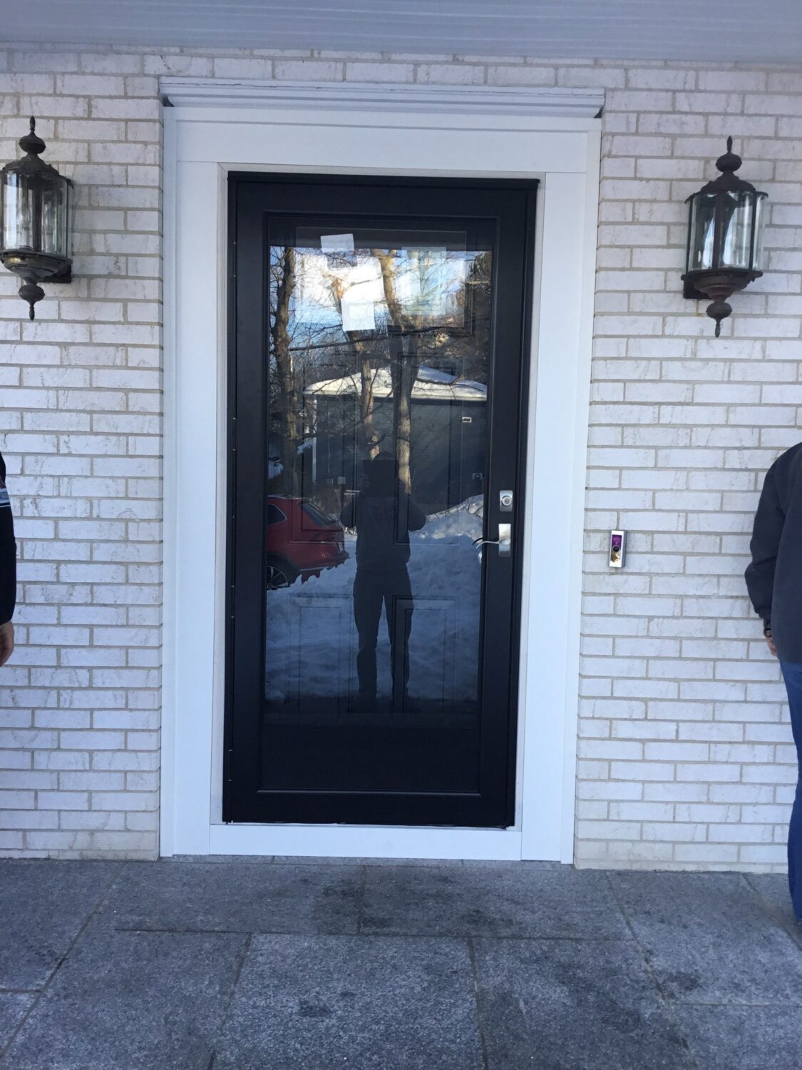 A black-framed glass door set in a white brick wall, flanked by two lantern-style light fixtures. Two people are partially visible on either side, and the door’s glass reflects the snowy outdoor scene.