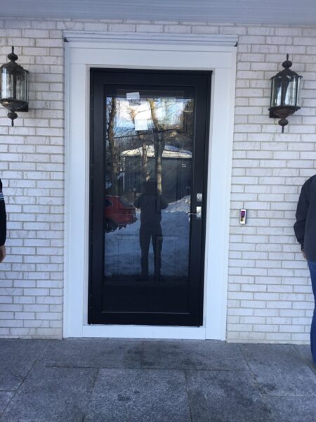 A black-framed glass door set in a white brick wall, flanked by two lantern-style light fixtures. Two people are partially visible on either side, and the door’s glass reflects the snowy outdoor scene.