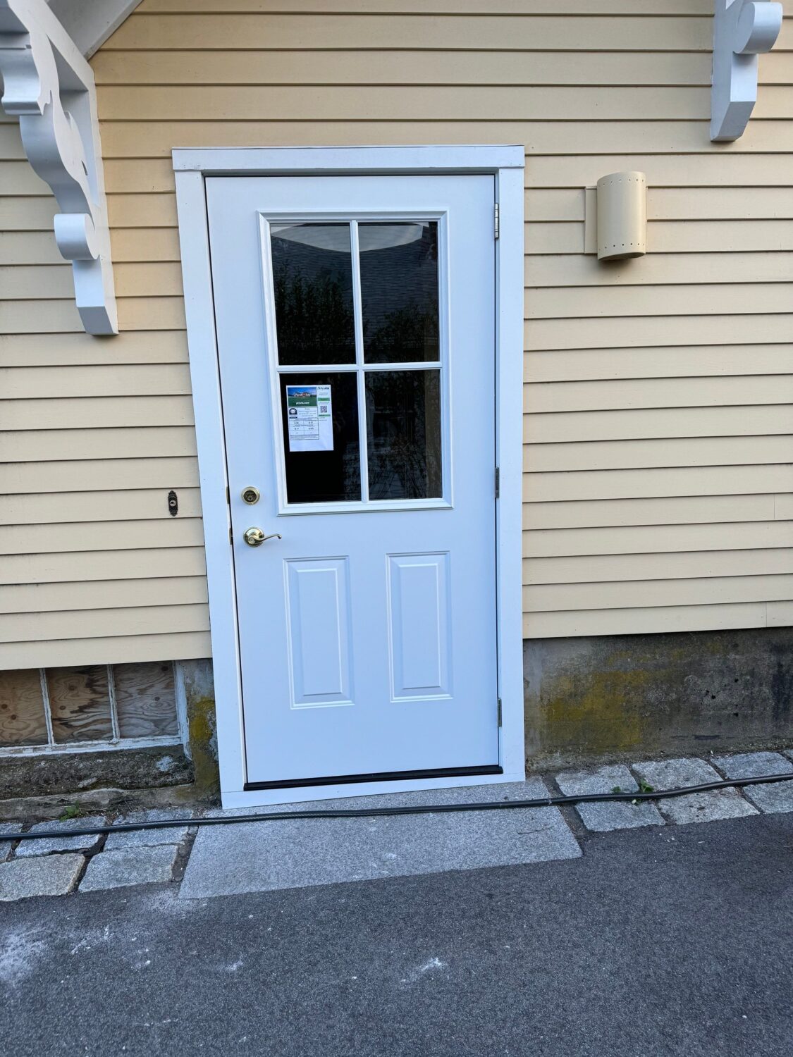 A white exterior door with a windowpane is set in a yellow house with horizontal siding. There is a notice or sticker on the window, and the ground is paved with stones and asphalt.