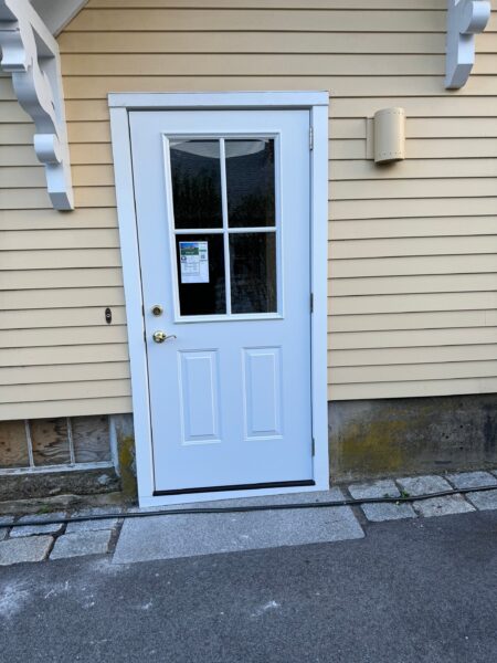 A white exterior door with a windowpane is set in a yellow house with horizontal siding. There is a notice or sticker on the window, and the ground is paved with stones and asphalt.