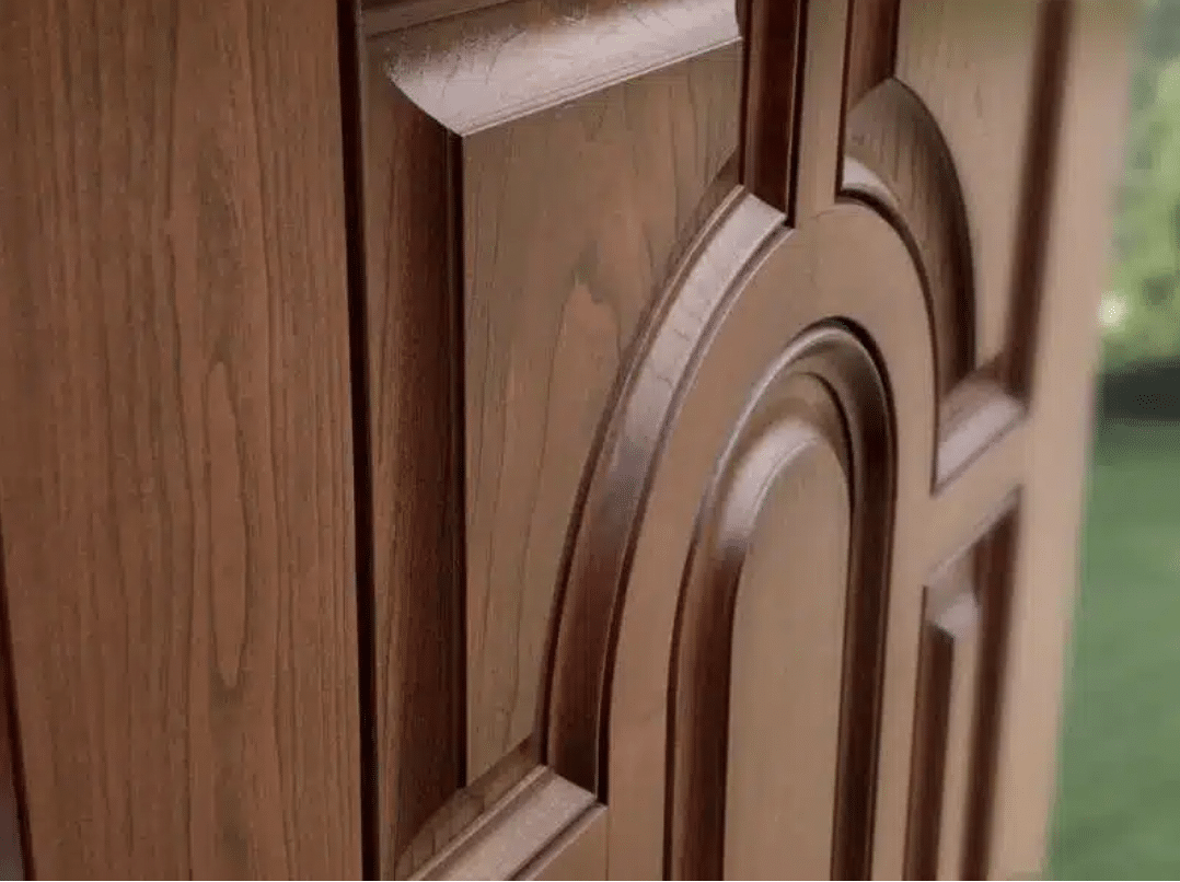 Close-up of a wooden door with an intricate design featuring curved and angular patterns. The wood has a rich, natural finish, echoing the elegance of classic windows. Blurred greenery is visible in the background.