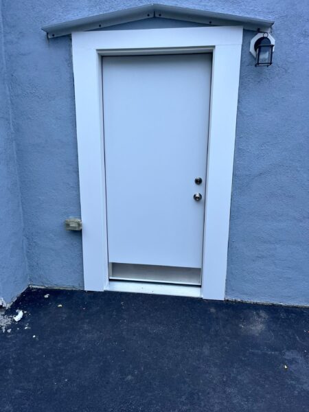 A small white door on a light blue wall with a small roof overhang. There is a silver doorknob and a second knob above it. A black sconce light is mounted on the wall to the right of the door. The ground is dark pavement.
