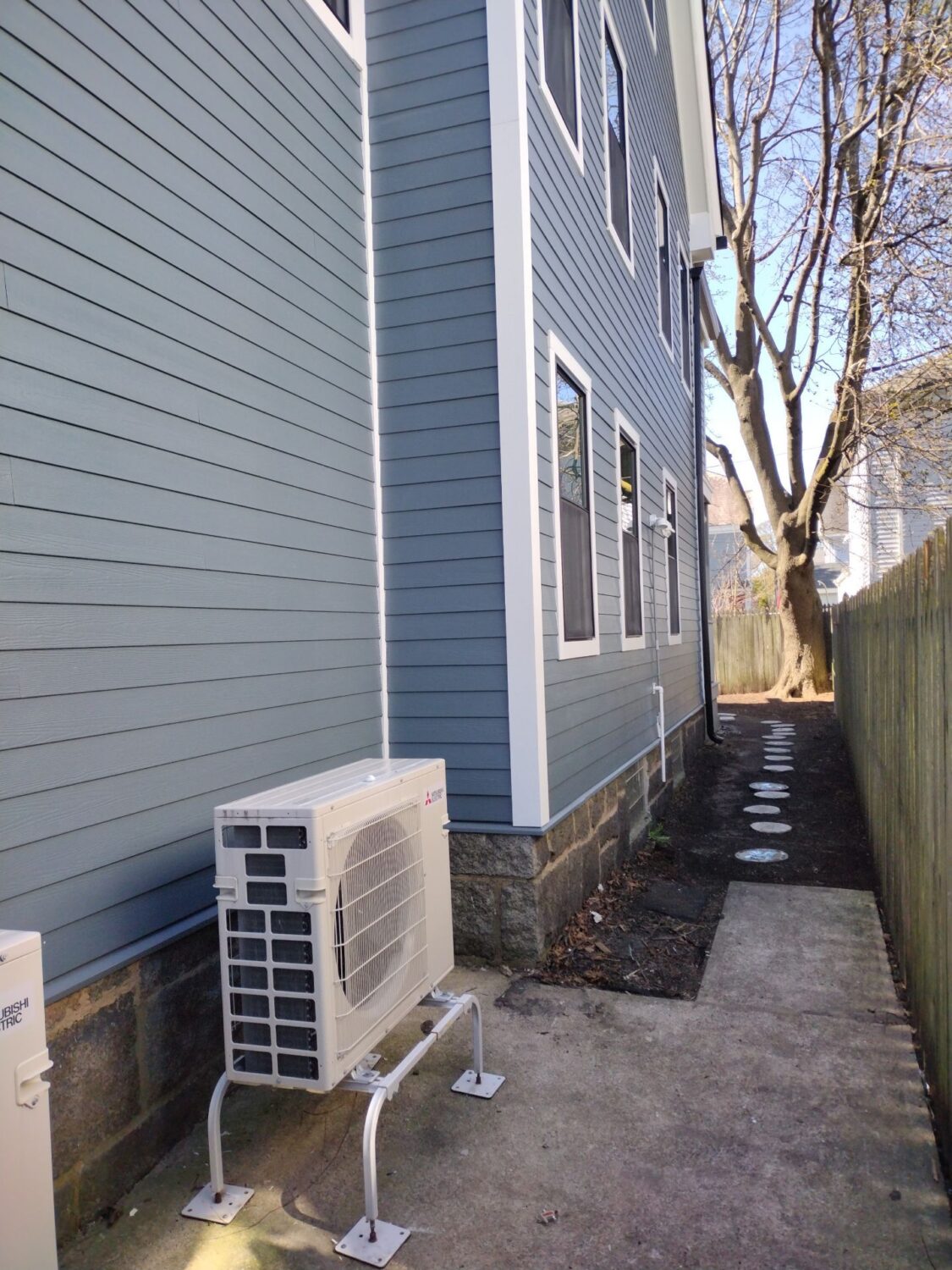 A narrow concrete path runs beside a blue house with white trim; an outdoor air conditioning unit is mounted along the house, and a tree stands at the end of the path next to a wooden fence.