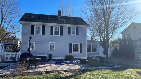 A two-story light gray house with black shutters, a brick chimney, and a fenced backyard with outdoor patio furniture and leafless trees under a clear blue sky.