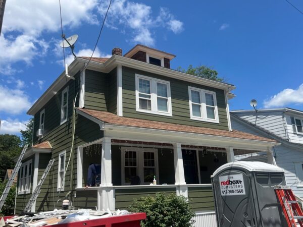 Two-story green house with brown roof, white trim, and multiple windows. Satellite dish on top. Ladders, a red dumpster, and a portable toilet are in front. Clear blue sky with a few clouds.