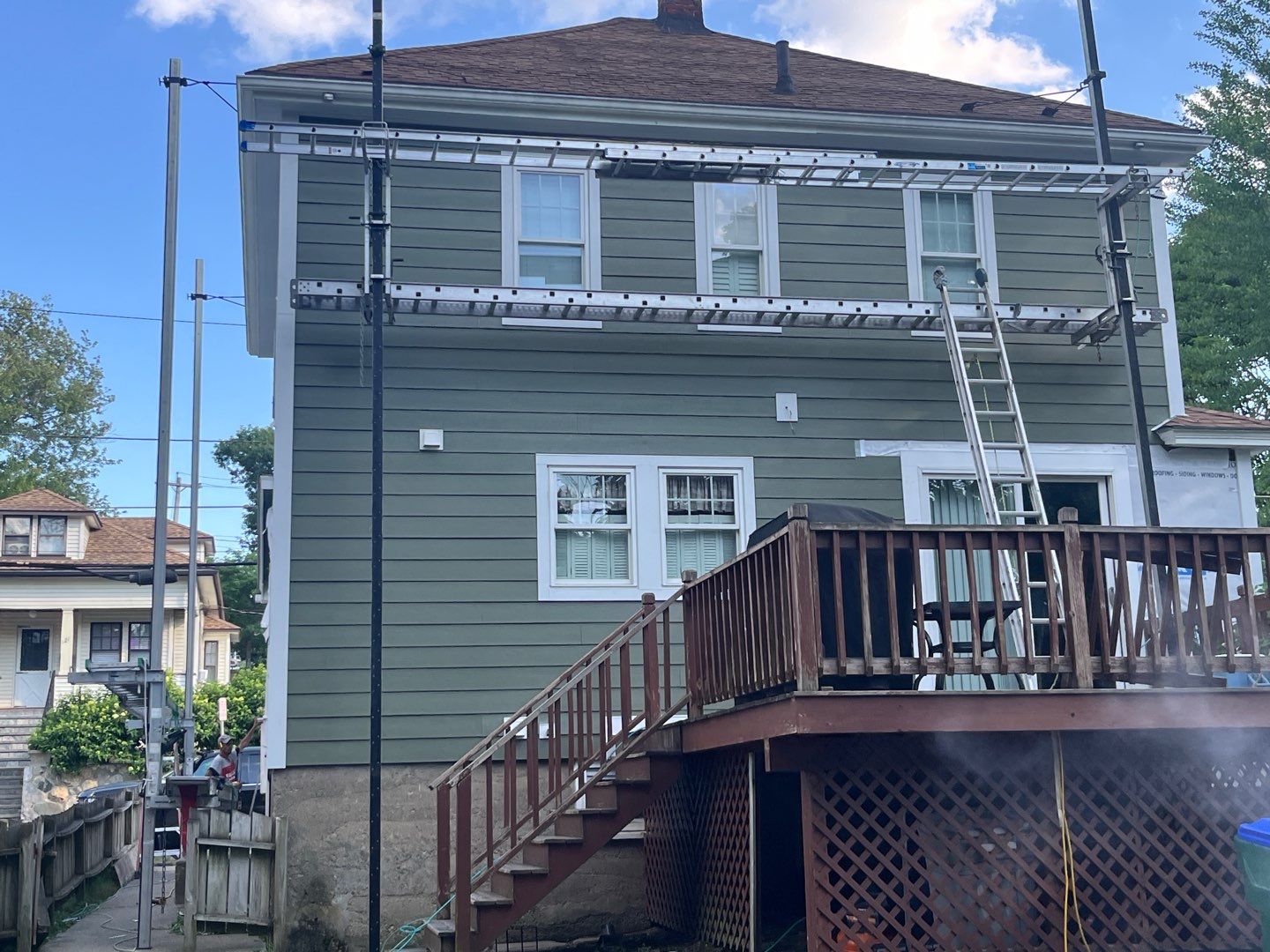 A green two-story house with scaffolding and a ladder set up against the upper level. A wooden deck with stairs leads to the backyard. The sky is partly cloudy and nearby houses are visible.