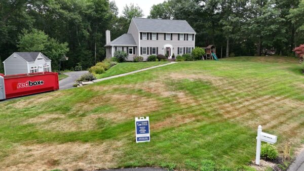 A large, gray, two-story house with a porch and a playset in the backyard is surrounded by trees. A sign on the lawn indicates a sale or event, and a red storage container is on the driveway. The lawn has dry patches.