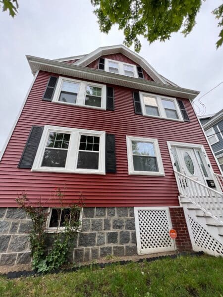 Red three-story house with black shutters, white trim, and a stone foundation. A small garden is under the lower windows. A white lattice panel is by the front entrance with a partially visible security sign on the grass.