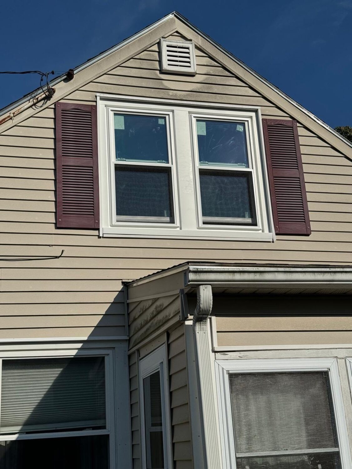 A beige house with a triangular roof and a pair of windows on the upper floor, framed by dark shutters. Below, a smaller window is partly shaded by a roof extension. The house shows some weathering.