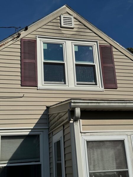 A beige house with a triangular roof and a pair of windows on the upper floor, framed by dark shutters. Below, a smaller window is partly shaded by a roof extension. The house shows some weathering.