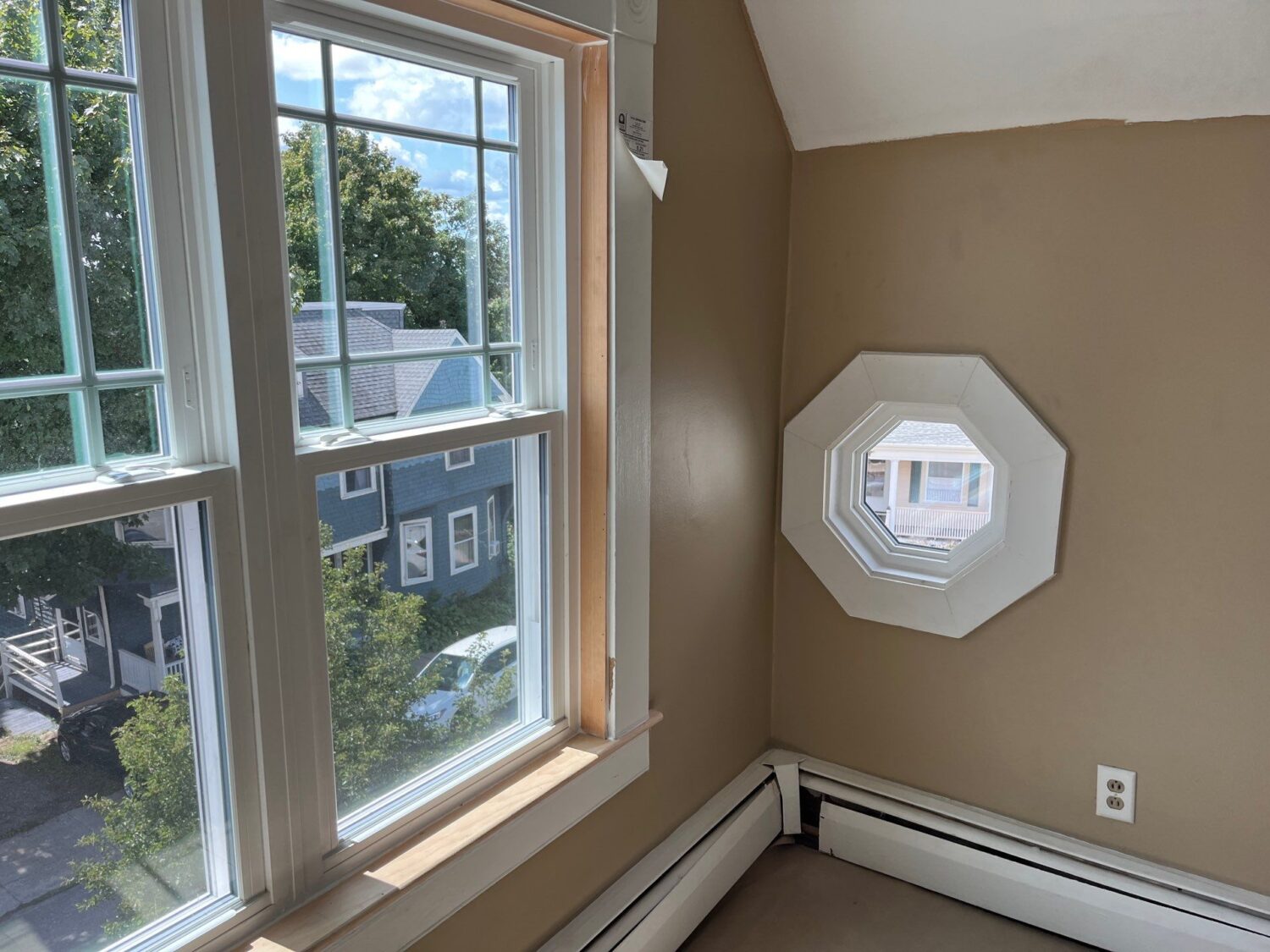 A sunlit room with beige walls features a large window on the left, showing a view of houses and trees outside. To the right is an octagonal window. Below the windows are baseboard heaters.