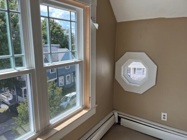 A sunlit room with beige walls features a large window on the left, showing a view of houses and trees outside. To the right is an octagonal window. Below the windows are baseboard heaters.