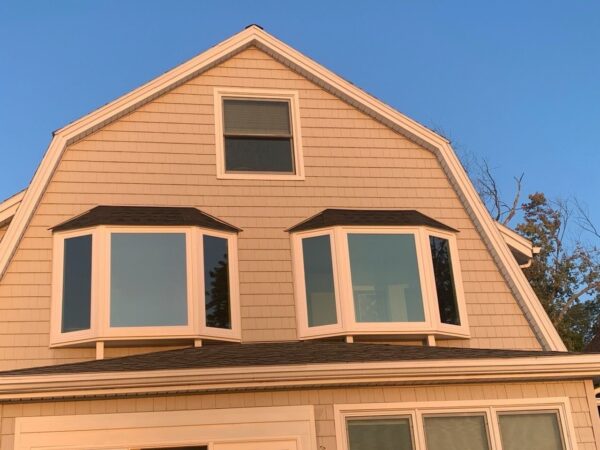 A house with light brown shingle siding and a steep gable roof appears illuminated by a warm sunset. It has two large bay windows on the second floor and one smaller window on the third floor. Clear blue sky in the background.