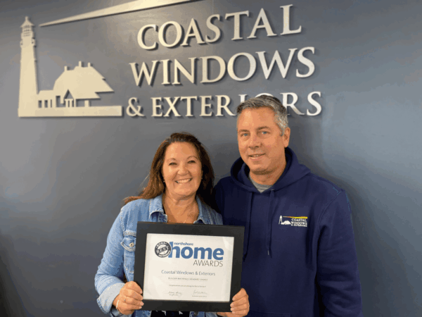 Two people smiling and holding a Home Awards certificate in front of a wall with the Coastal Windows & Exteriors logo and lighthouse graphic, celebrating recognition among the best Boston home improvement companies.