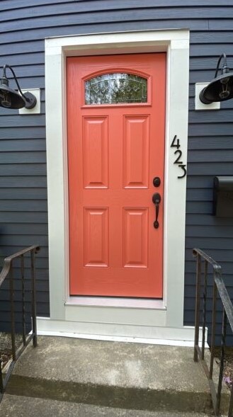 A coral-colored front door with a small window is set in a white frame on a dark blue house. Entry Doors Wakefield MA style is showcased by the vertically mounted house number 423 and two black wall lanterns flanking the entry.
