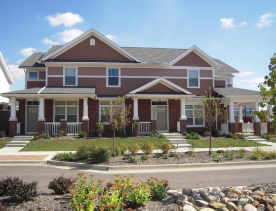 A two-story duplex with Hardie Siding in Wakefield MA features brown siding, white trim, and covered front porches. Young trees and shrubs line the sidewalk and street, beneath a clear blue sky.