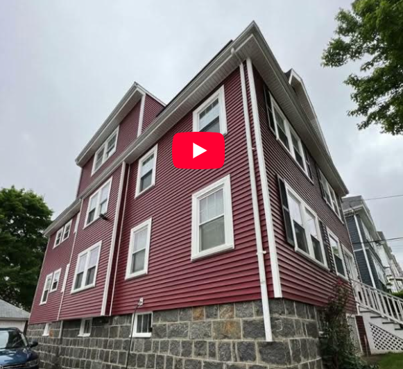 A three-story house with red siding and white trim sits on a stone foundation under a cloudy sky, with green trees and a parked blue car partially visible—just the kind of home featured by Coastal Windows & Exteriors Founder in the Boston Herald.