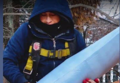 A person wearing a blue hooded jacket, yellow safety harness, and gloves is holding a large, light blue cylindrical object outdoors on a cold dayāpossibly part of the renovation industry, as seen in recent Wrentham news.