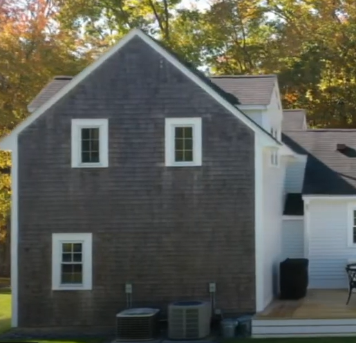 A house with gray wooden siding and white trim, featured in the Hopedale Town News Feature for HIP 200 List, with three side windows, outdoor air conditioning units, autumn trees behind, and sunlight casting shadows on the home.