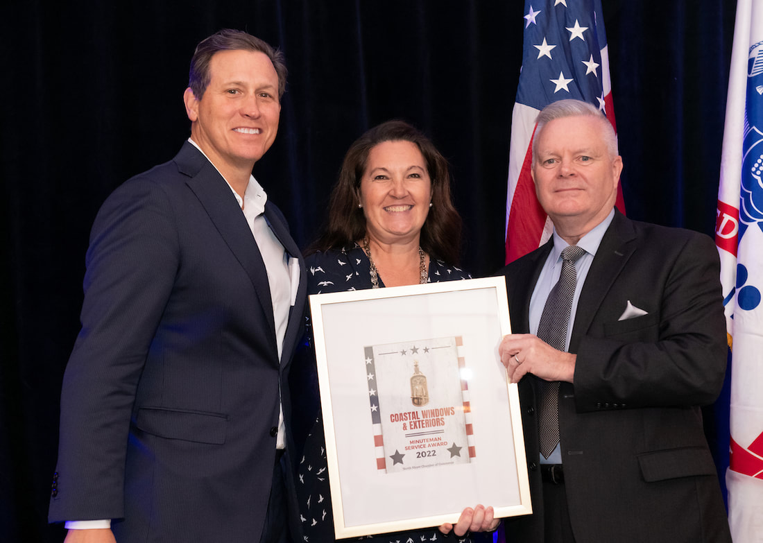Three people in formal attire smile while holding a framed award, possibly recognizing one of the best Salisbury home improvement companies. An American flag and another flag in the background suggest an official or celebratory event.