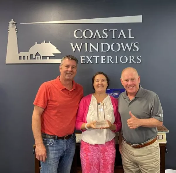 Three people stand smiling in front of a Coastal Windows Exteriors sign; the woman in the center holds a glass award, showcasing why they’re recognized among the best Ashland home improvement companies. The two men beside her give a thumbs up.
