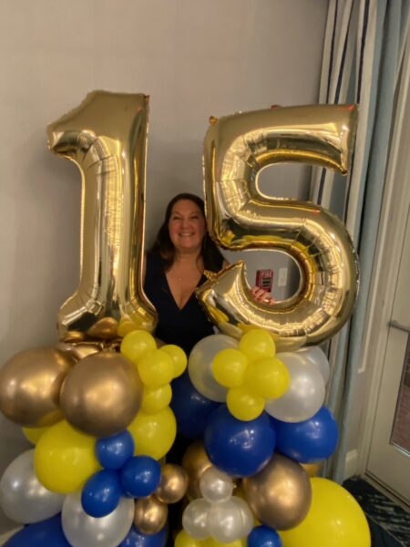 A smiling woman stands between large gold balloons shaped as the number 15, surrounded by blue, yellow, and gold balloon decorations—celebrating with one of the best Attleboro home improvement companies.
